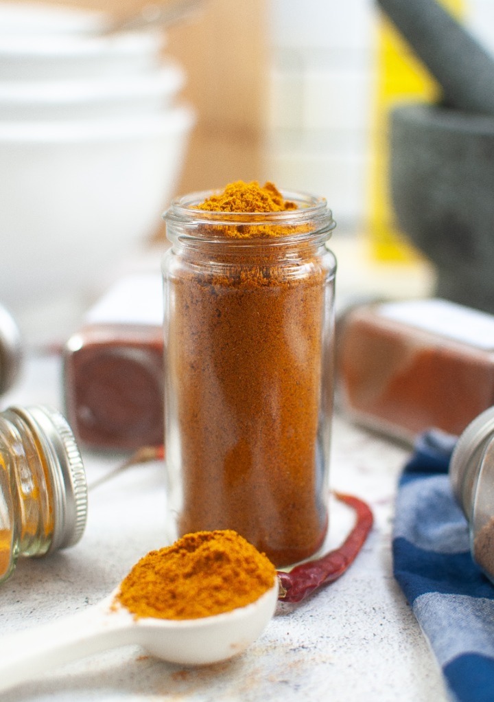 Homemade curry powder in a clear herb jar with a scoop on the side. 