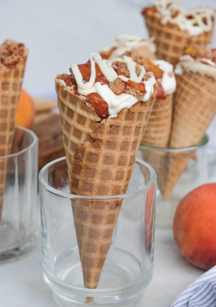 Waffle cone filled with peach cobbler held up in a clear glass.