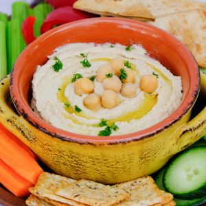 Creamy hummus in a yellow bowl with orange rim surrounded by vegetables.
