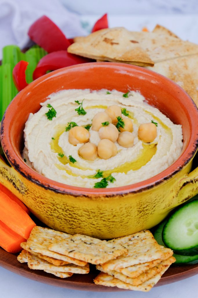 Homemade hummus in a yellow dish with crackers and vegetables on the side. 