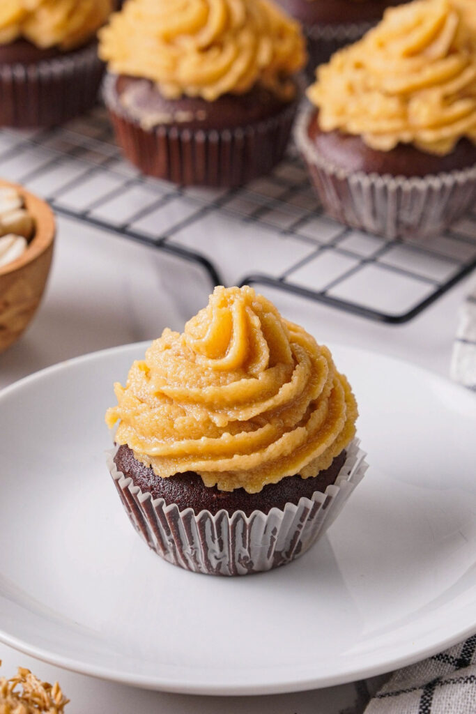 A chocolate cupcake with peanut butter frosting on a white serving dish with more cupcakes on a baking rack. 