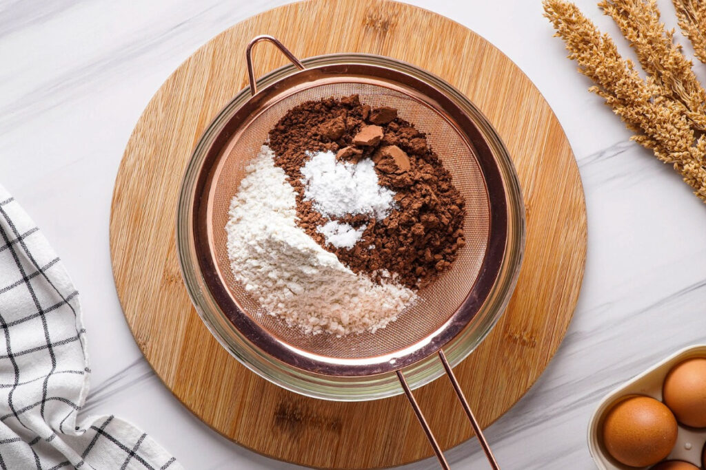 Sifting cocoa, flour, salt, and baking soda over a clear bowl to make cupcakes. 