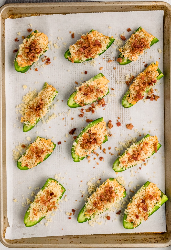 Jalapenos stuffed with sausage on a baking sheet topped with bacon.