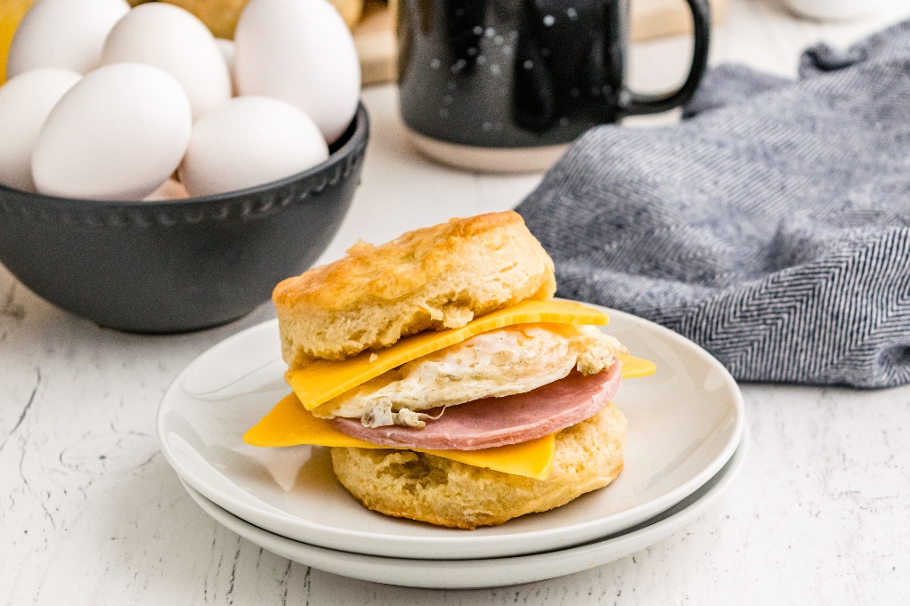 A biscuit sandwich with fresh eggs in the background and a cup of coffee.