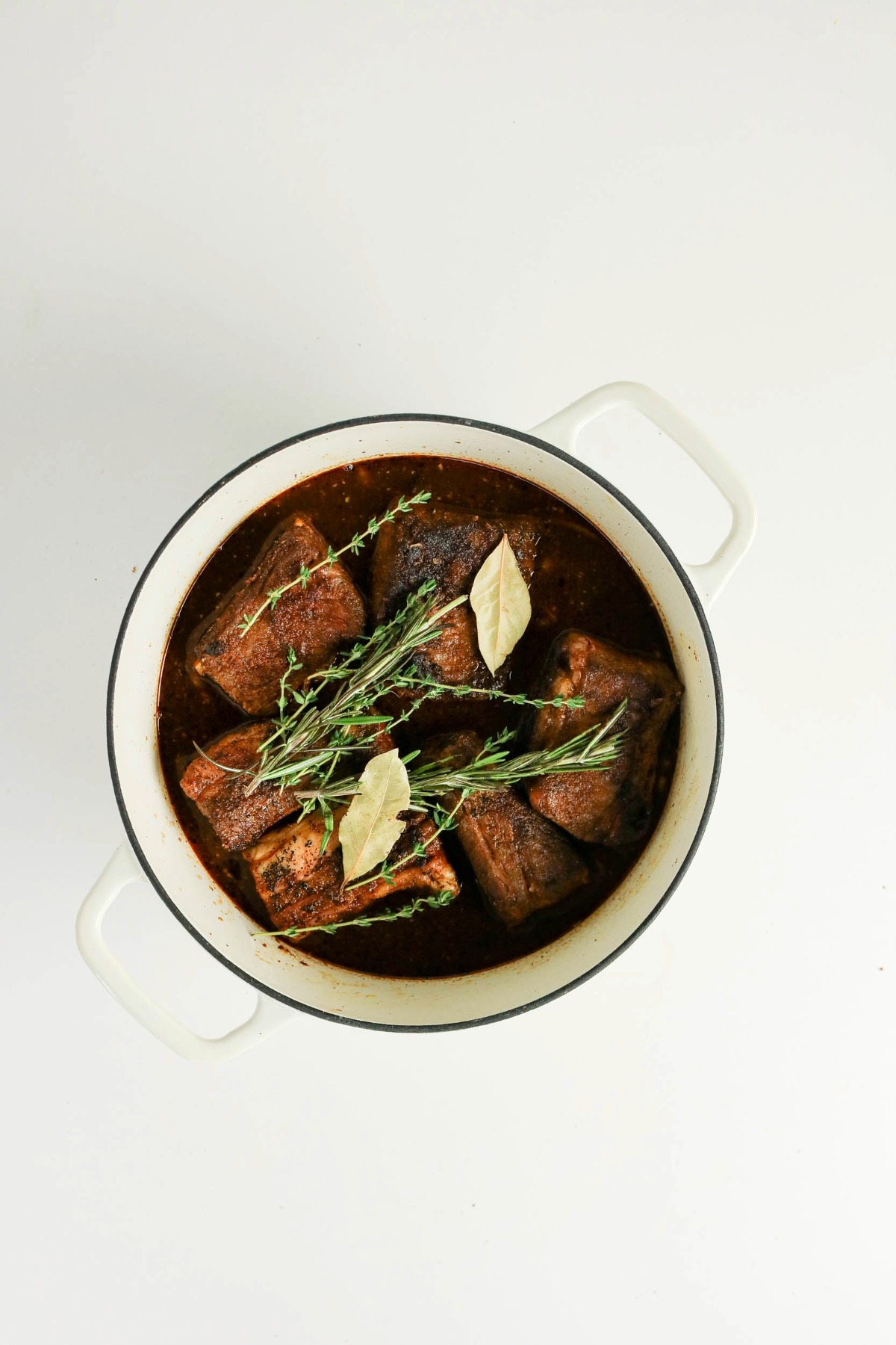 Short ribs in a Dutch oven with fresh herbs before covering and cooking.