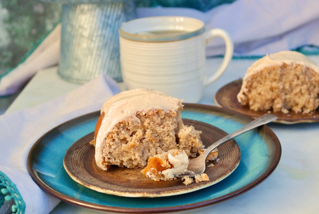 A slice of spice cake topped with icing with a mug on the side ready to be enjoyed. 