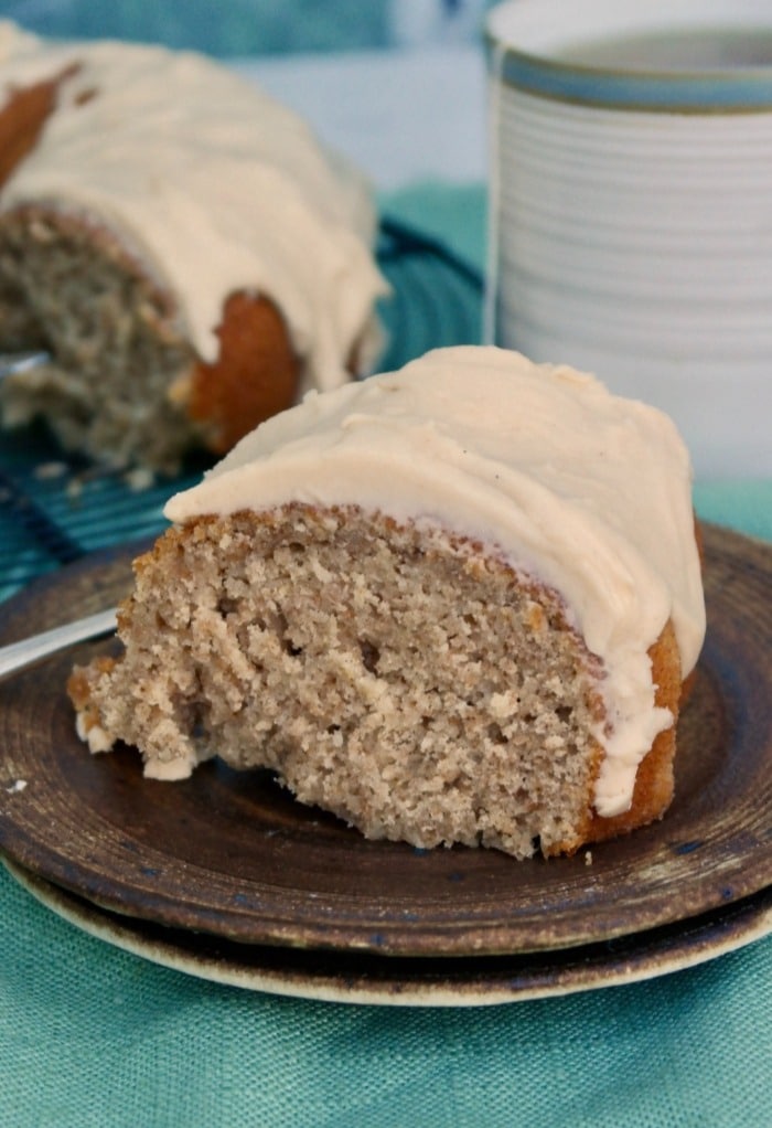 A slice of spice cake on a brown serving place with a cup of coffee in the background. 