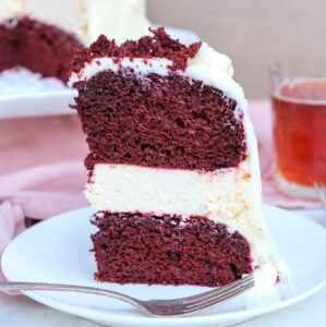 A close view of a three layered red velvet cheesecake cake on a white plate ready to eat.