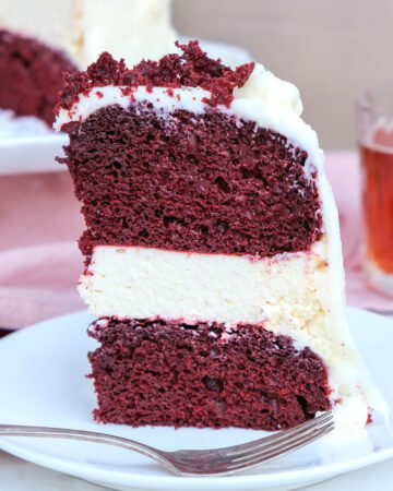 A close view of a three layered red velvet cheesecake cake on a white plate ready to eat.