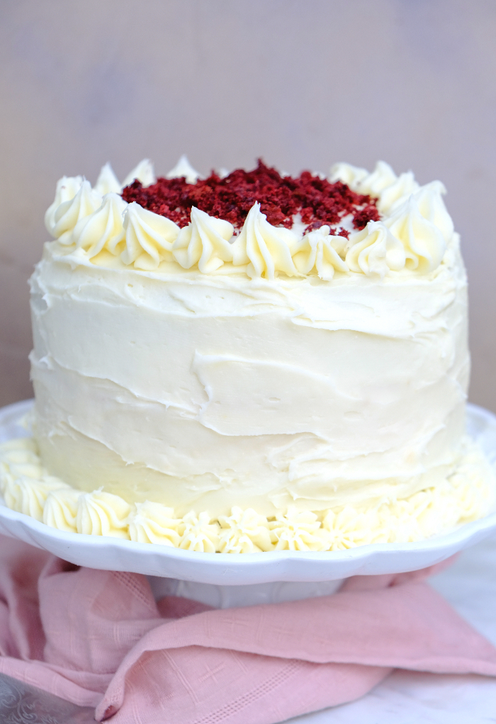 A whole red velvet cake on a cake platter with cream cheese frosting.