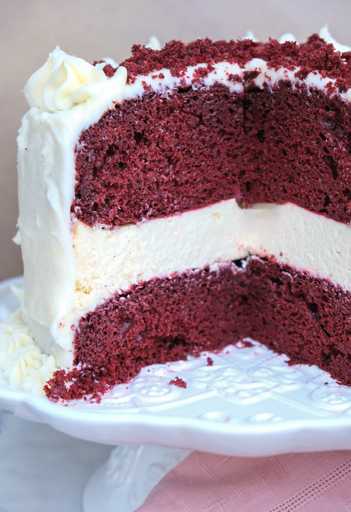 A slice of a triple layer red velvet and cheesecake cake on a white cake platter. 