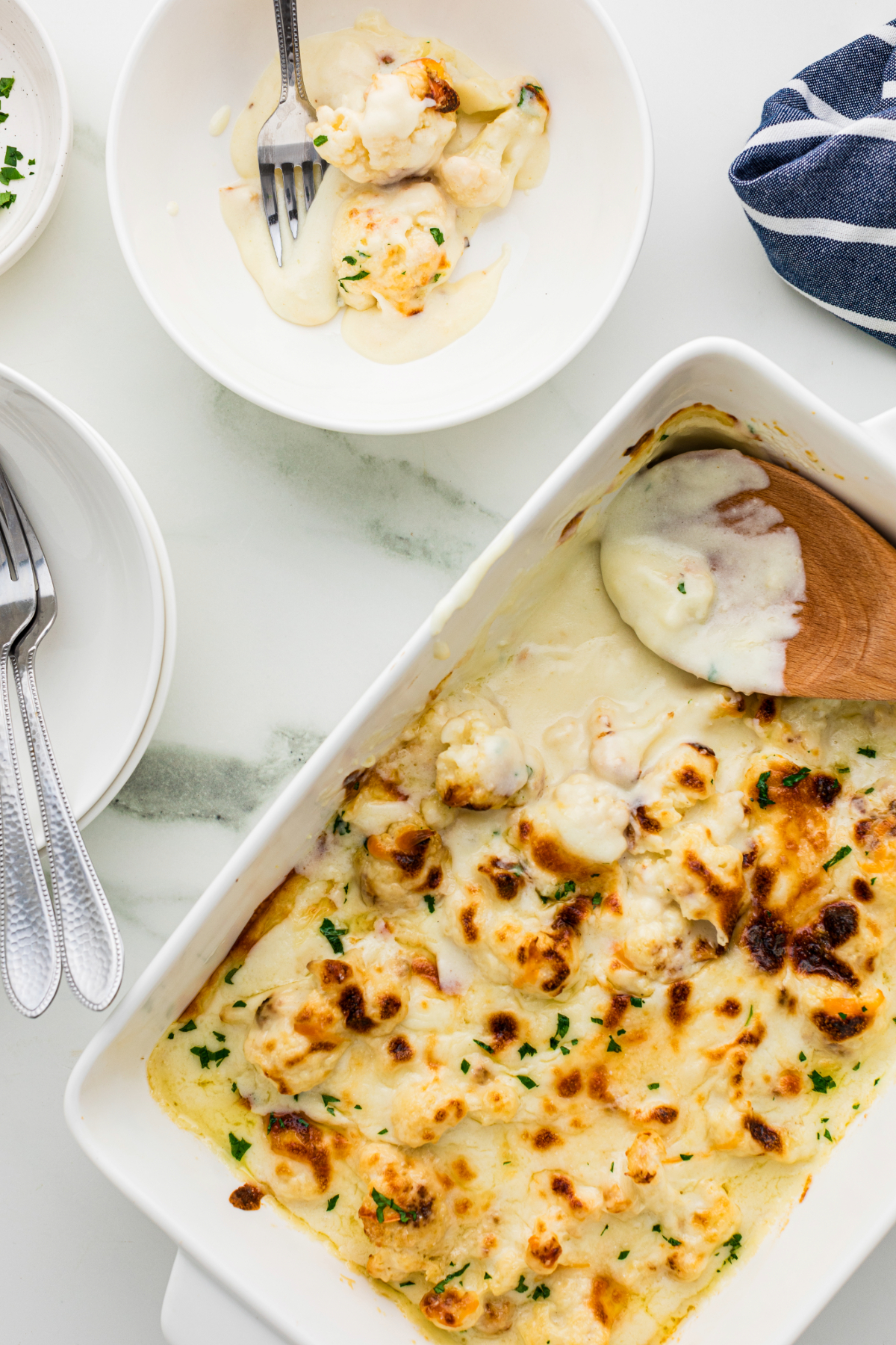 Overhead view of cheesy cauliflower white casserole dish with a wooden spoon serving a scoop on a white dish. 