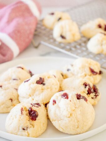 A white plate full of cranberry coconut cookies with cookies cooling on a backing rack in the background.