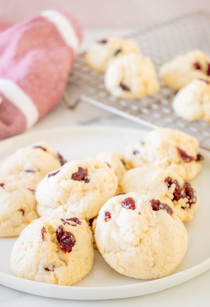 A white plate full of cranberry coconut cookies with cookies cooling on a backing rack in the background.