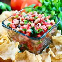 Texas caviar dip in a clear bowl with scoop chips around the bowl on a wood platter.