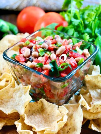 Texas caviar dip in a clear bowl with scoop chips around the bowl on a wood platter.