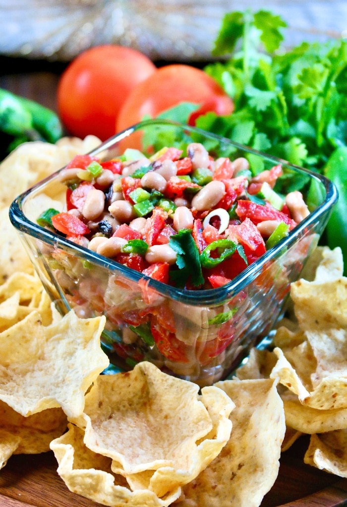 Texas caviar dip in a clear bowl with scoop chips around the bowl on a wood platter. 