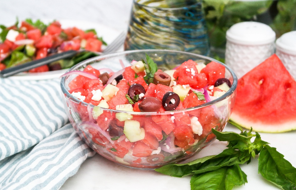 Watermelon salad in a clear mixing bowl with olives and feta cheese. 