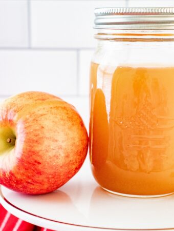 A clear mason jar with homemade apple pie moonshine on a white cake plate with a fresh apple on the side.