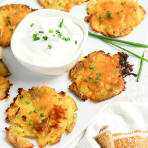 A small bowl of sour cream topped with chives next to a white plate of smashed potatoes.