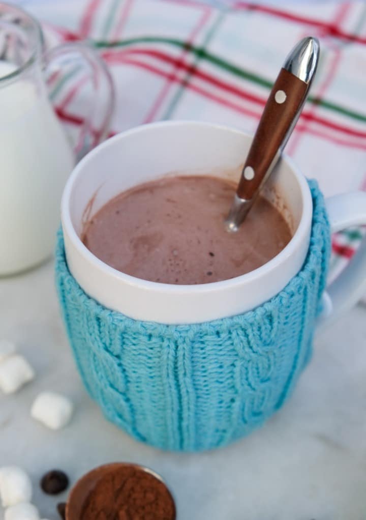 Hot cocoa in a white mug with a teal cozy around the cup. 
