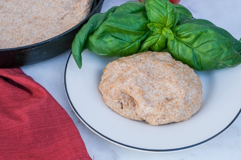 Homemade pizza dough on a white plate with fresh basil on the side. 