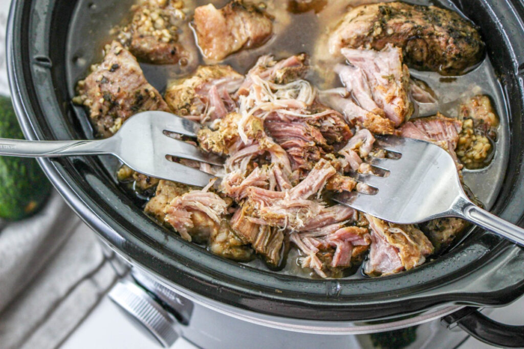 Slow cooker with cooked carnitas shredding with two forks. 