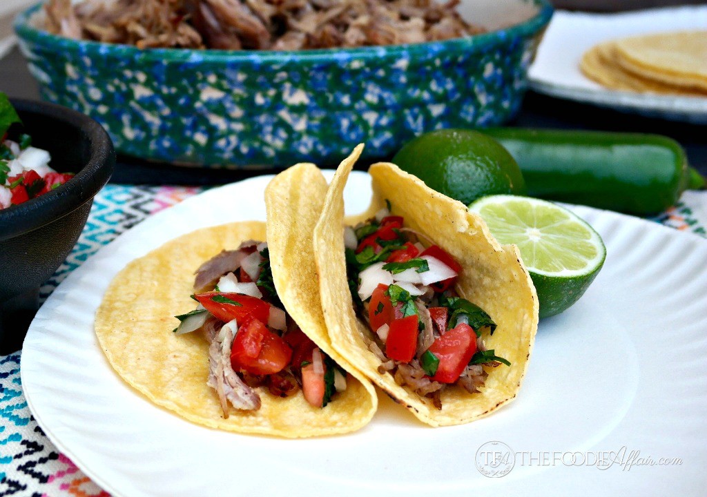 two slow Cooker Pork Carnitas wrapped in corn tortillas on a white plate with slice of lime on the side