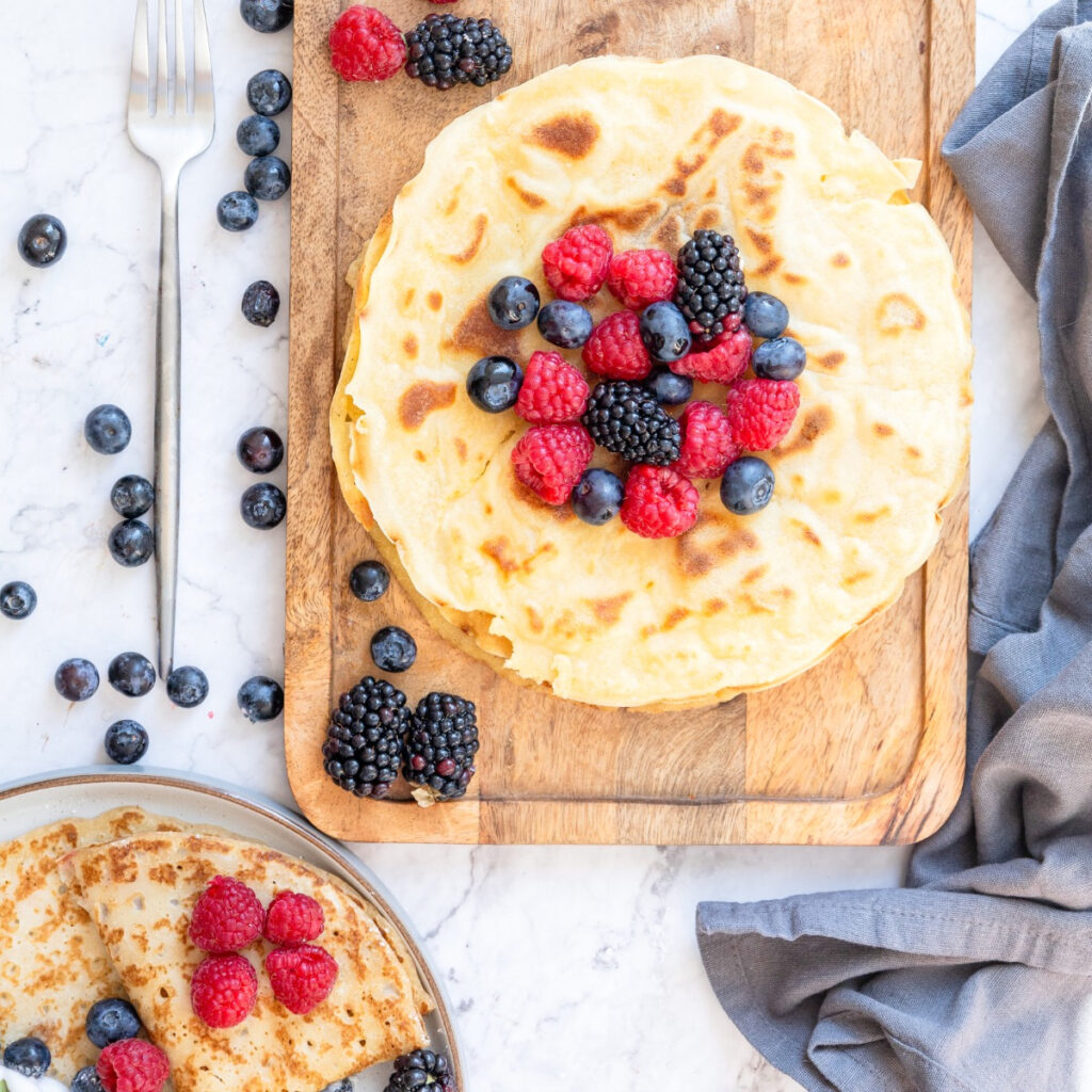A stack of fresh cooked crepes on a cutting board with fresh fruit on top of the stack.