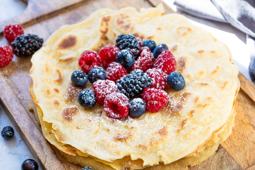 A stack of cooked crepes on a cutting board topped with fruit.