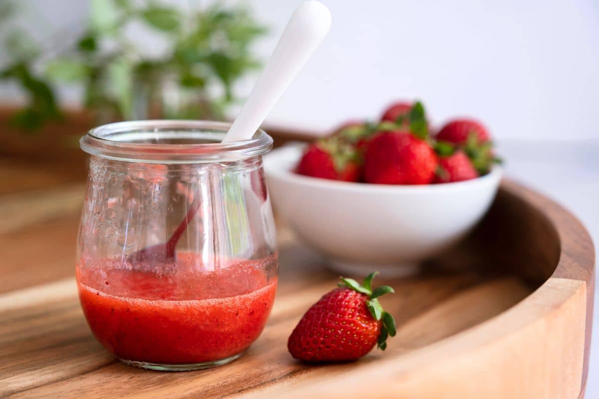 Strawberry puree in a small glass jar with a spoon on a wooden tray.