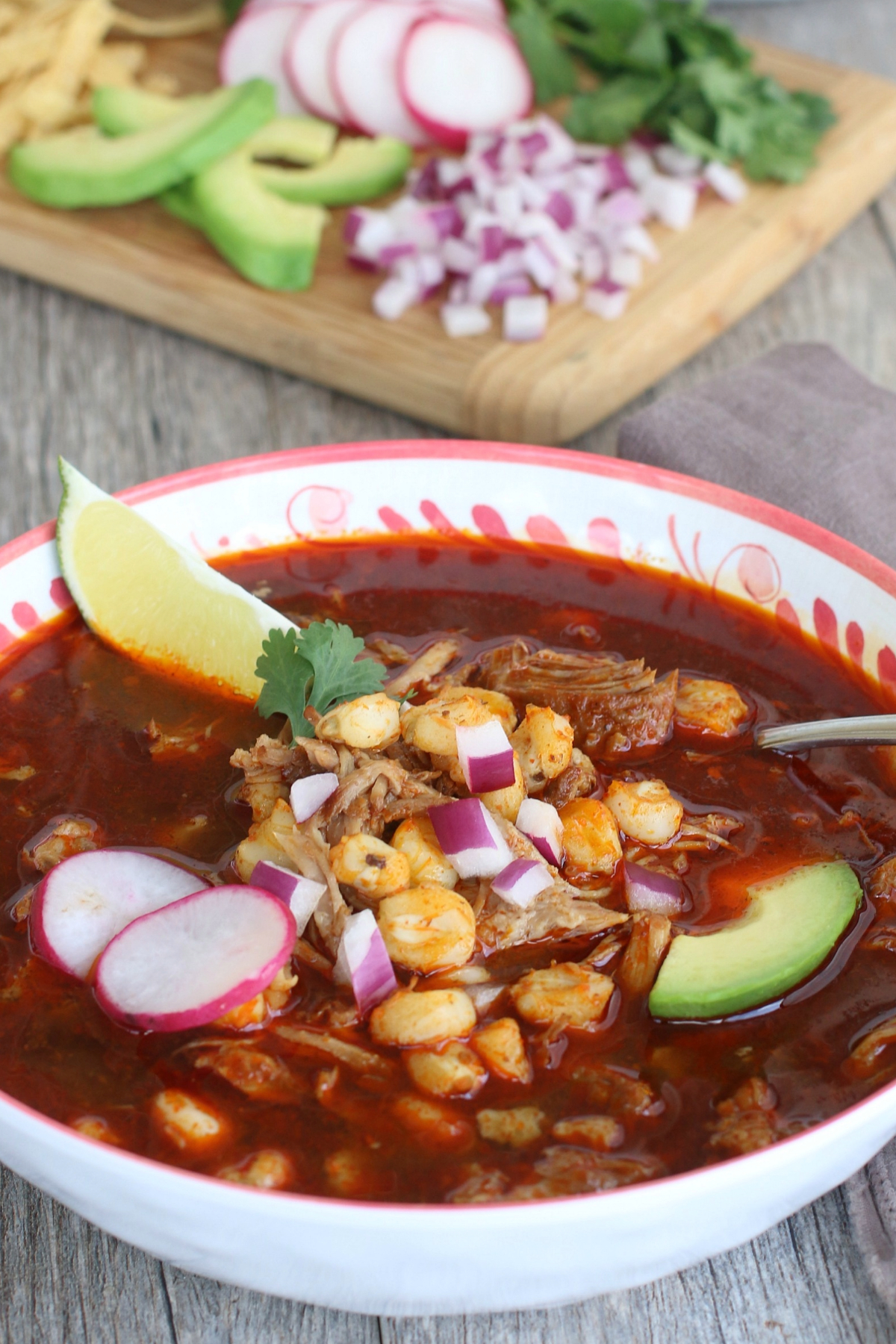 A bowl with pozole with red chili sauce topped with radish, avocado and a slice of lime.