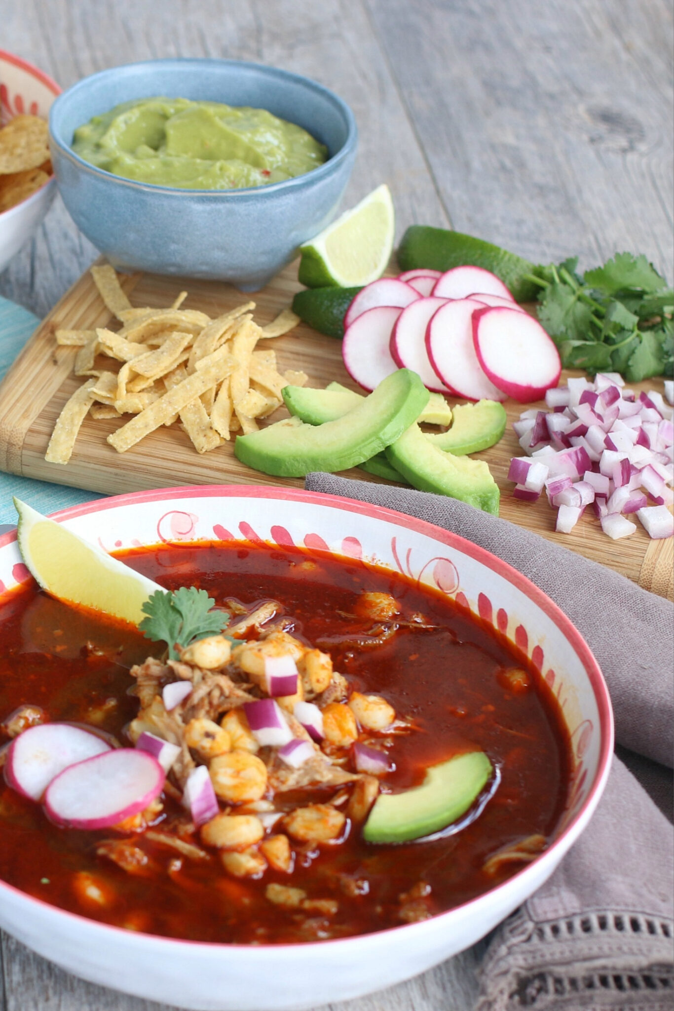 A bowl of pozole with toppings in the background on a wooden cutting board.