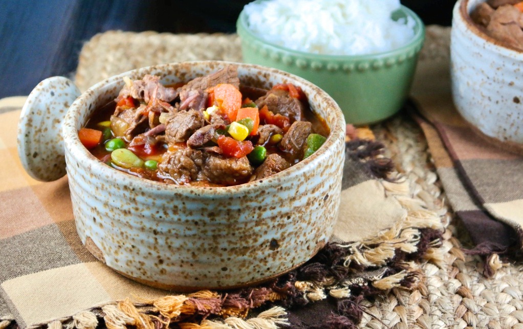 A soup bowl filled with beef and vegetables with a side dish of rice. 