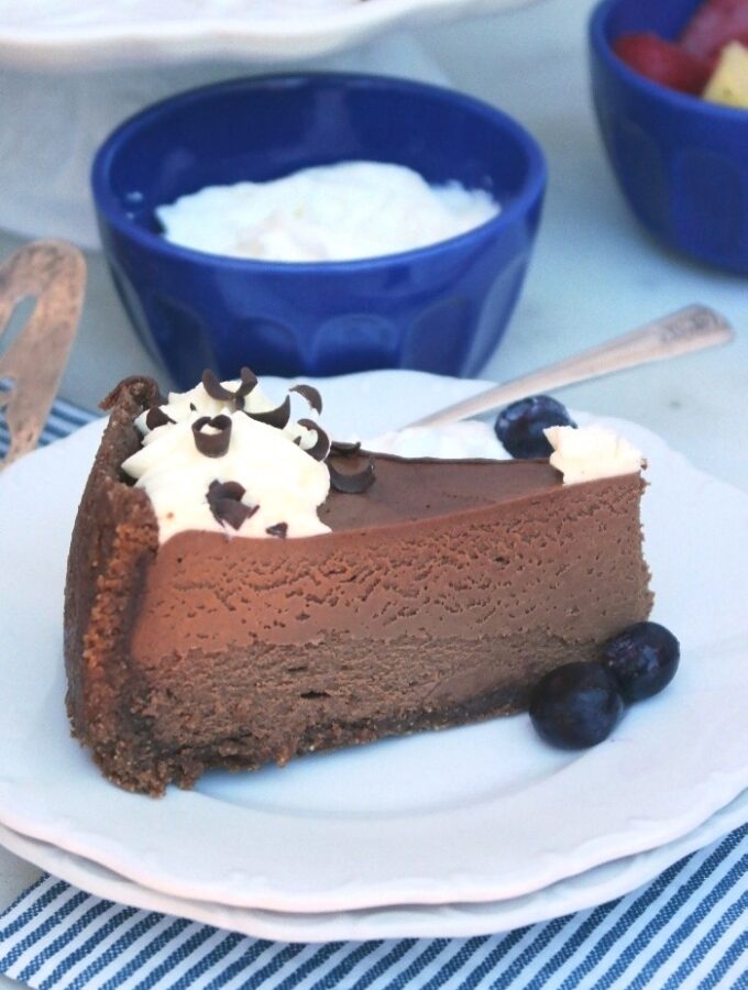 Large slice of chocolate cheesecake on a white plate with a blue bowl of whipped cream in the back.