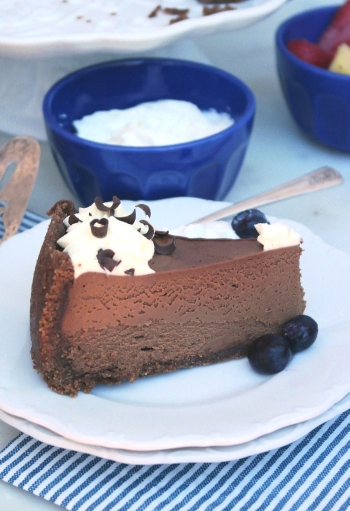 Large slice of chocolate cheesecake on a white plate with a blue bowl of whipped cream in the back.