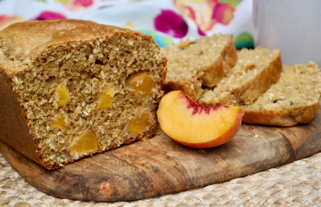 A loaf of peach bread on a cutting board with a slice of fresh peach on the side