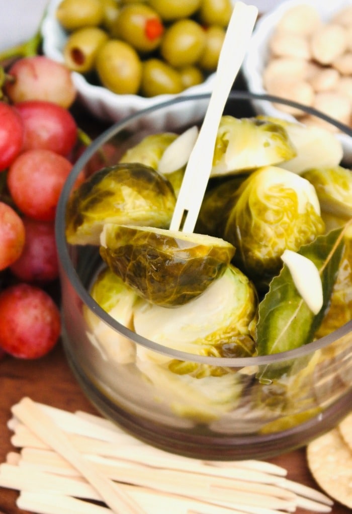 Pickled brussels sprouts in a glass bowl surrounded by grapes and olives to be enjoyed. 