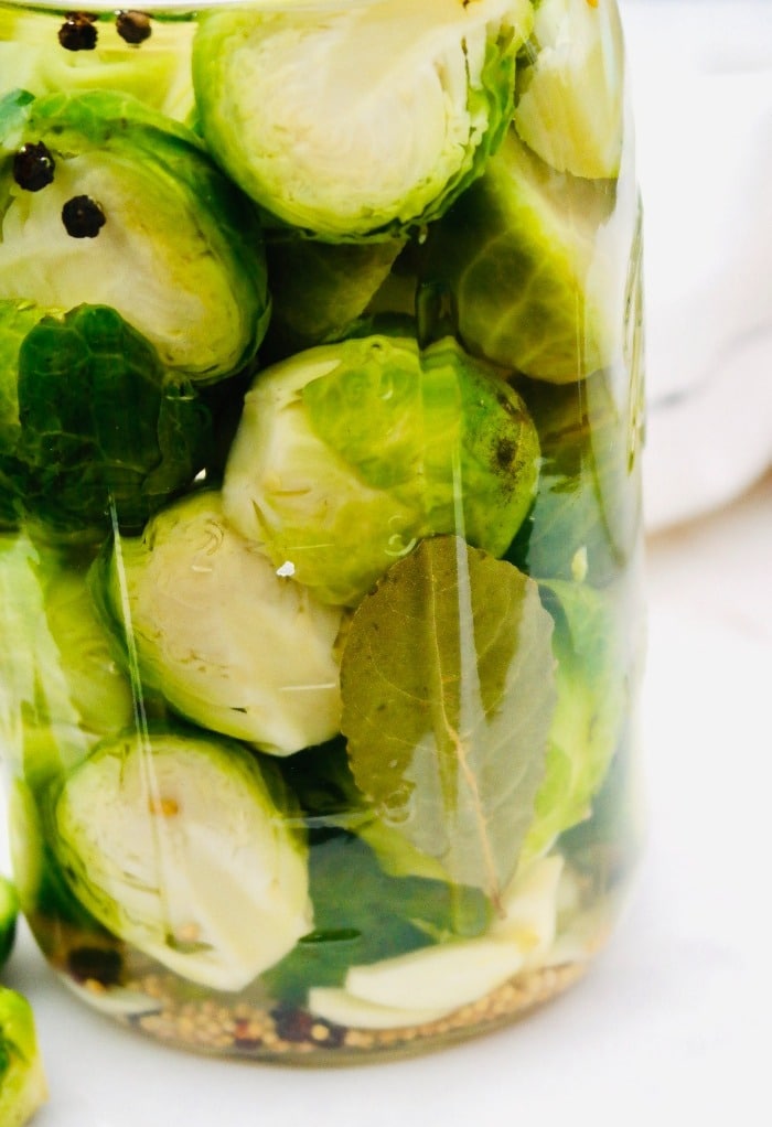 Pickled brussels sprouts in a clear glass mason jar with a bay leaf.