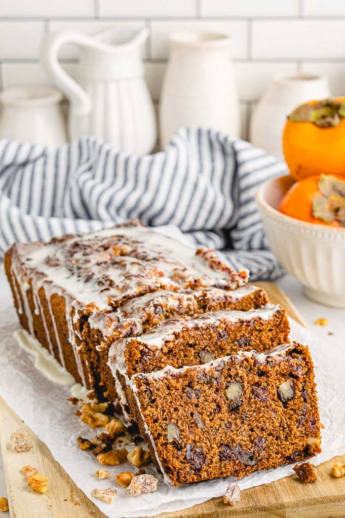 Sliced persimmon bread with nuts and topped with a glaze on a cutting board.