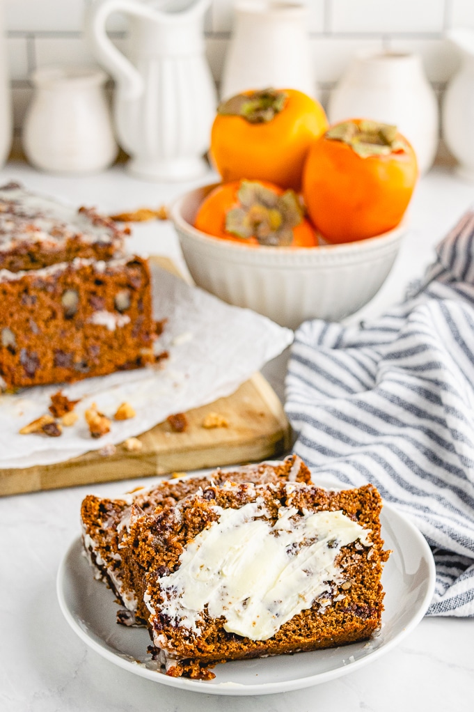 Sliced persimmon bread on a white plate with fresh persimmons in the background. 