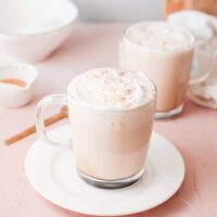 Clear mug on a white plate filled with caramel latte topped with whipped cream.