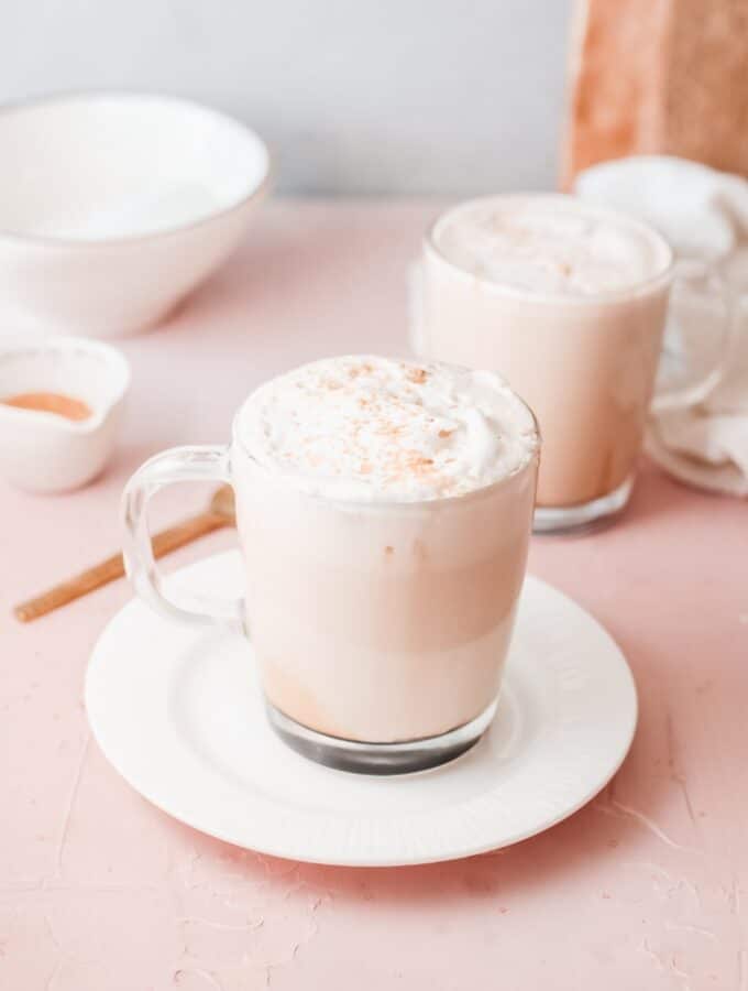 Clear mug on a white plate filled with caramel latte topped with whipped cream.