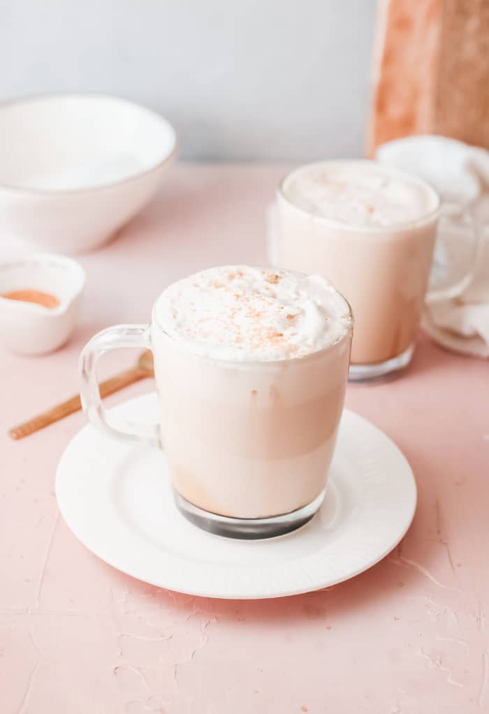 Clear mug on a white plate filled with caramel latte topped with whipped cream.