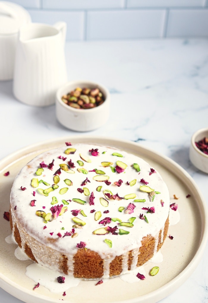 Persian love cake topped with pistachios and rose petals on a white serving plate.  