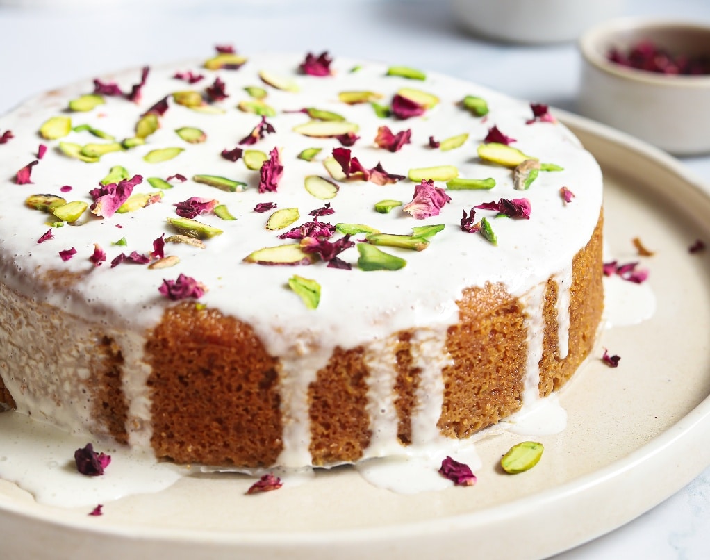 Decorated Persian love cake topped with rose petals and pistachios on a cream serving cake plate. 