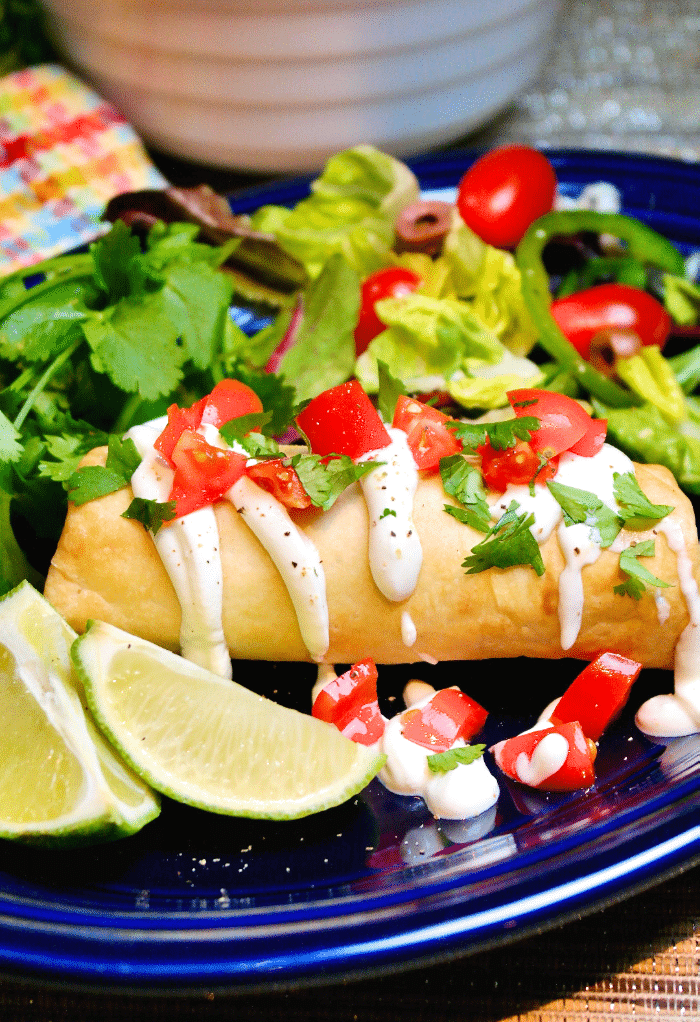 Chimichanga topped with tomatoes and sour cream on a blue plate ready to eat.