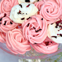 A glass cake platter filled with pink and white rose meringue cookies topped with dried flowers.