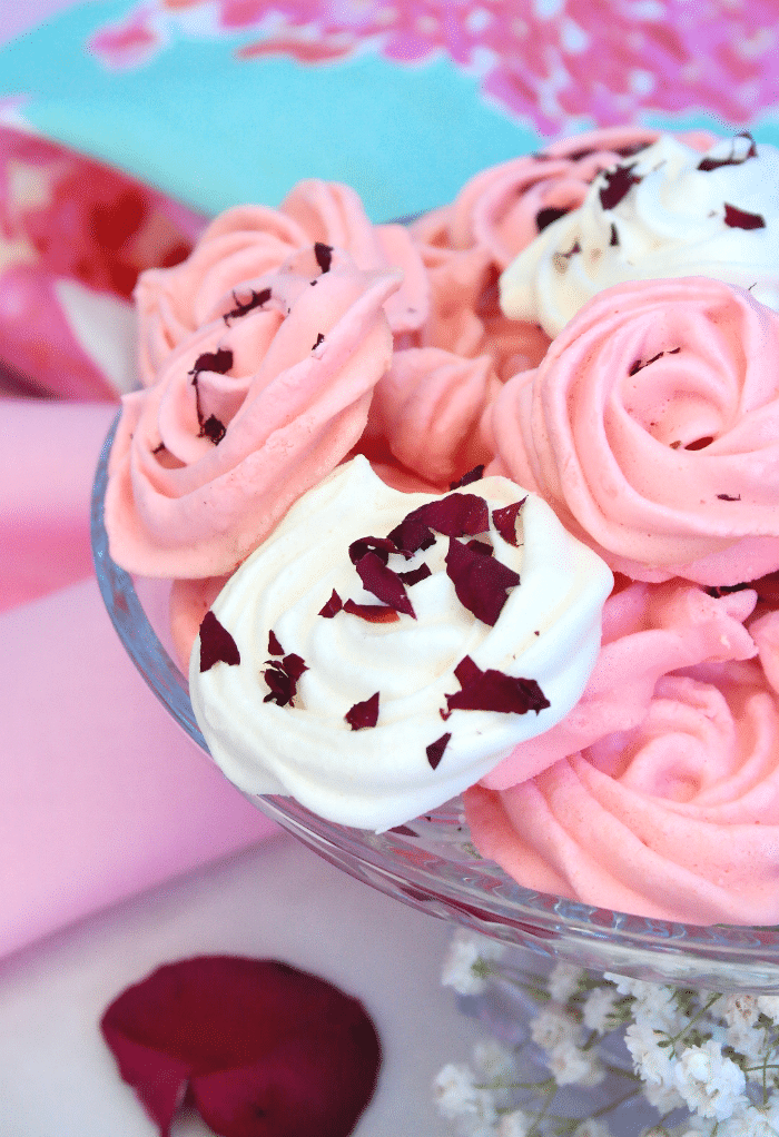 Pink and white meringue cookies on a clear cake stand with dried roses on top. 