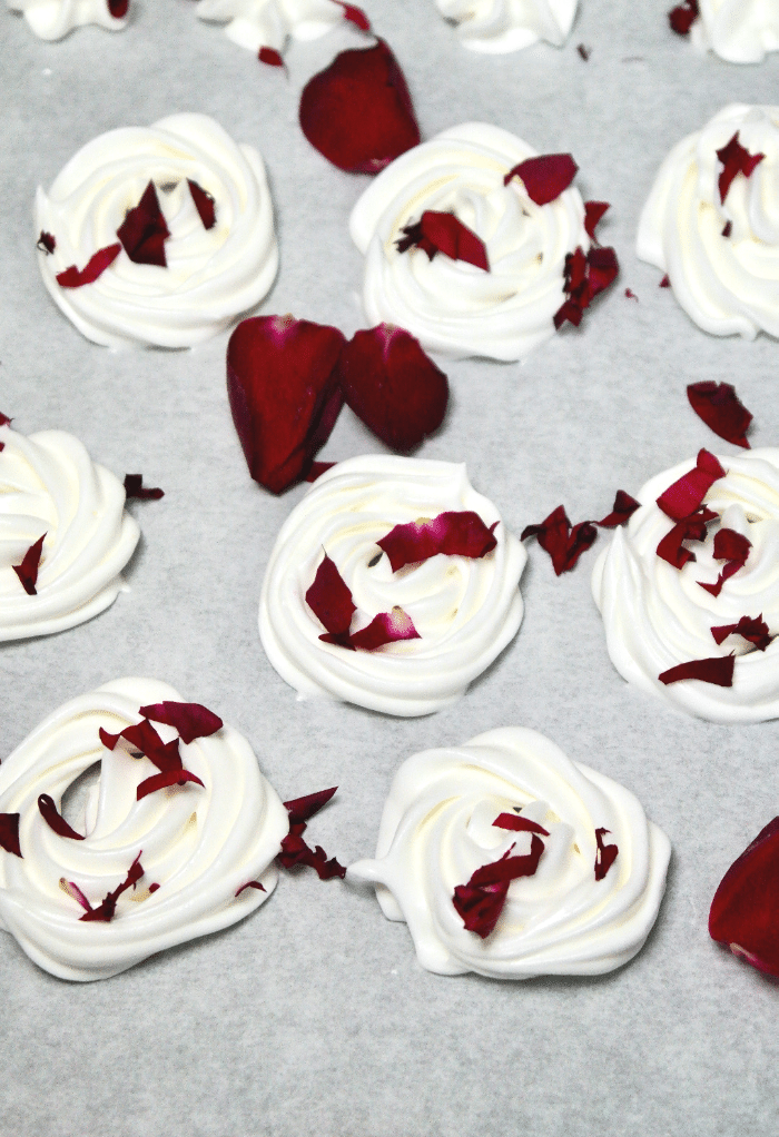 White rose meringue cookies on parchment paper before baking topped with rose petals. 